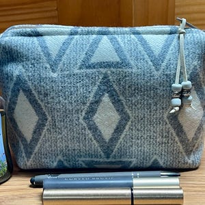 Puede incluir: Un neceser rectangular con estampado azul claro y blanco, con cierre de cremallera y una peque&ntilde;a borla. La bolsa presenta un dise&ntilde;o geom&eacute;trico de diamantes y tri&aacute;ngulos. Una botella de gel de ducha Country Chic y un bol&iacute;grafo est&aacute;n al lado de la bolsa.