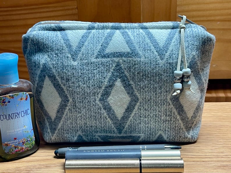 Puede incluir: Un neceser rectangular con estampado azul claro y blanco, con cierre de cremallera y una peque&ntilde;a borla. La bolsa presenta un dise&ntilde;o geom&eacute;trico de diamantes y tri&aacute;ngulos. Una botella de gel de ducha Country Chic y un bol&iacute;grafo est&aacute;n al lado de la bolsa.