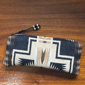 May include: A rectangular pencil case with a Southwestern geometric pattern in black, cream, and brown. The case has a zipper and a beaded pull. The case is made of a woven fabric.
