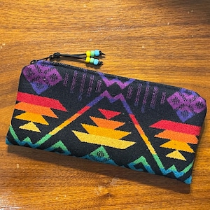 May include: A black zippered pouch with a vibrant geometric pattern in rainbow colors. The design features zigzags and mountain-like shapes. The zipper pull has small yellow, blue, and black beads.