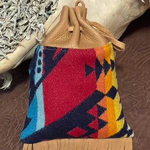 May include: A small, drawstring pouch with a colorful geometric pattern in red, blue, yellow, and orange. The top is made of tan leather, and the bottom has a fringe detail. The pouch is likely used for carrying small items.
