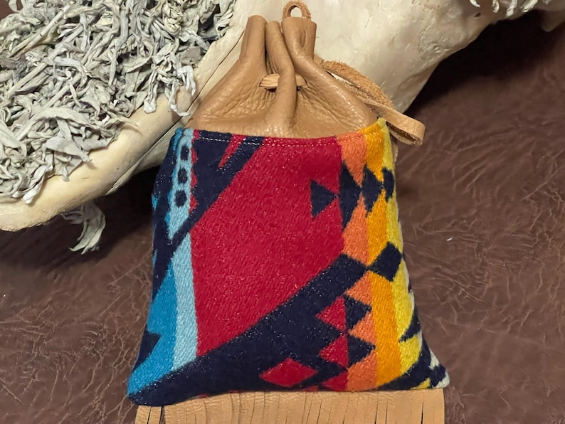 May include: A small, drawstring pouch with a colorful geometric pattern in red, blue, yellow, and orange. The top is made of tan leather, and the bottom has a fringe detail. The pouch is likely used for carrying small items.