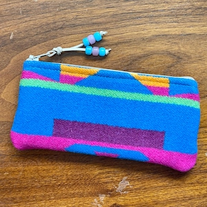 May include: A rectangular zippered pouch made of woven fabric featuring a vibrant geometric pattern in shades of bright blue, hot pink, purple, orange, and lime green. The zipper pull is accented with four small, alternating light blue and lavender beads.