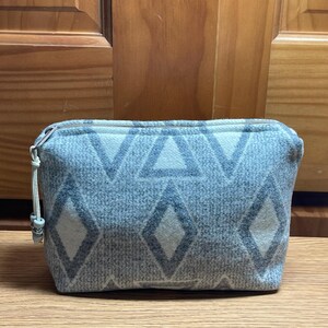 Puede incluir: Un neceser rectangular con cremallera y un estampado geom&eacute;trico de diamantes y tri&aacute;ngulos grises sobre un fondo azul claro. El neceser tiene una cremallera clara y un peque&ntilde;o tirador decorativo.