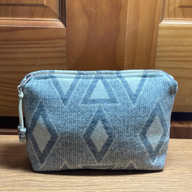 Puede incluir: Un neceser rectangular con cremallera y un estampado geom&eacute;trico de diamantes y tri&aacute;ngulos grises sobre un fondo azul claro. El neceser tiene una cremallera clara y un peque&ntilde;o tirador decorativo.