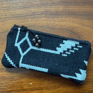 May include: A rectangular black and white patterned pencil case with a zipper closure. The case features a geometric design in white against a black background. The zipper pull has decorative beads.