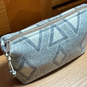 Puede incluir: Un estuche con cremallera con estampado gris y blanco. El estuche tiene un dise&ntilde;o geom&eacute;trico de diamantes y tri&aacute;ngulos. La cremallera es de color marr&oacute;n claro y el tirador tiene un peque&ntilde;o adorno de cuentas. El estuche est&aacute; hecho de una tela suave y texturizada.