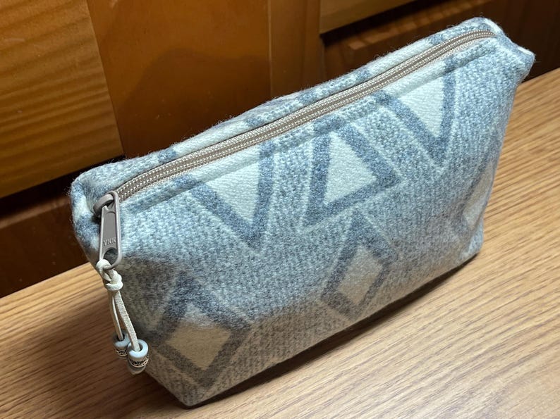 Puede incluir: Un estuche con cremallera con estampado gris y blanco. El estuche tiene un dise&ntilde;o geom&eacute;trico de diamantes y tri&aacute;ngulos. La cremallera es de color marr&oacute;n claro y el tirador tiene un peque&ntilde;o adorno de cuentas. El estuche est&aacute; hecho de una tela suave y texturizada.