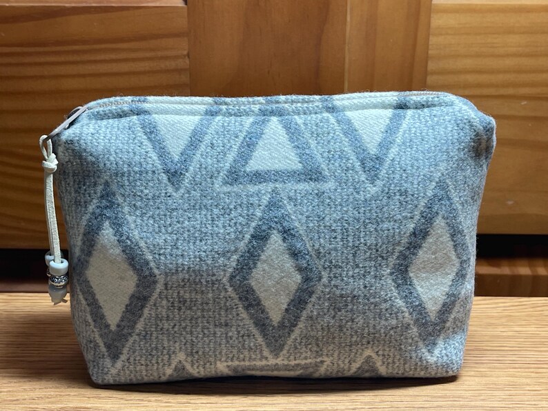 Puede incluir: Peque&ntilde;o neceser rectangular con cierre de cremallera. El neceser es de color azul claro con un patr&oacute;n repetido de diamantes y tri&aacute;ngulos grises. Un peque&ntilde;o tirador con cuentas est&aacute; unido a la cremallera.