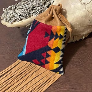 May include: A small, tan leather pouch with a drawstring closure. The pouch features a colorful, patterned fabric front with red, blue, yellow, and black geometric designs. Long, tan leather fringe hangs from the bottom.