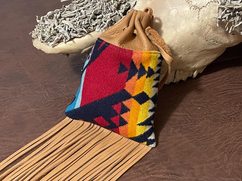 May include: A small, tan leather pouch with a drawstring closure. The pouch features a colorful, patterned fabric front with red, blue, yellow, and black geometric designs. Long, tan leather fringe hangs from the bottom.
