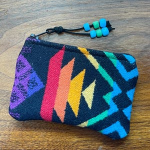 May include: A small, zippered pouch with a colorful geometric pattern in shades of purple, red, orange, yellow, green, and blue. The pouch has a black background and a black zipper pull with turquoise and green beads.