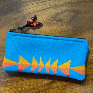 May include: A turquoise zippered pouch with a geometric design in orange and yellow. The pouch has a black zipper and a beaded pull with orange and brown beads. The pouch is rectangular and appears to be made of felt.