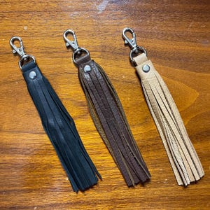 Könnte beinhalten: Drei Leder-Schlüsselanhänger mit Quasten in Schwarz, Braun und Beige. Jeder Schlüsselanhänger hat eine silberne Metallschaufel.