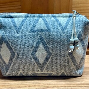 Puede incluir: Una bolsa rectangular con cremallera en tonos azules y blancos, con un patr&oacute;n geom&eacute;trico de diamantes y tri&aacute;ngulos. La bolsa tiene un tirador de cremallera azul claro con cuentas decorativas. La bolsa est&aacute; sobre una superficie de madera.