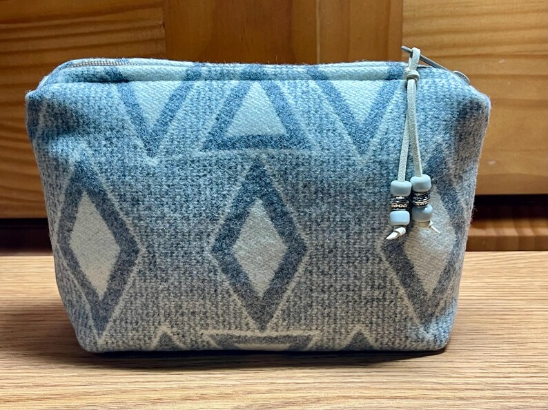 Puede incluir: Una bolsa rectangular con cremallera en tonos azules y blancos, con un patr&oacute;n geom&eacute;trico de diamantes y tri&aacute;ngulos. La bolsa tiene un tirador de cremallera azul claro con cuentas decorativas. La bolsa est&aacute; sobre una superficie de madera.