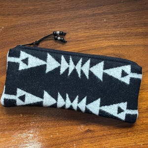 May include: A black pencil case with a repeating white arrow pattern. The case has a black zipper and a black cord with decorative beads. The case is made of a soft, textured material.