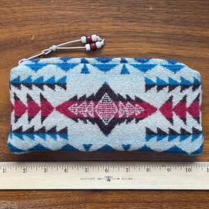 May include: A rectangular, zippered pouch with a geometric pattern in shades of blue, pink, and black. The pouch has a white cord with red and white beads attached to the zipper. A wooden ruler is placed below the pouch.