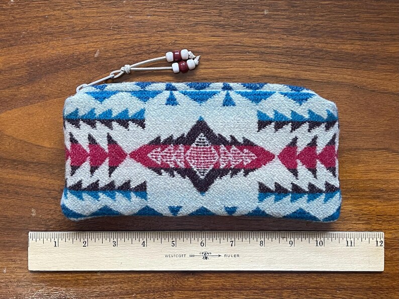 May include: A rectangular, zippered pouch with a geometric pattern in shades of blue, pink, and black. The pouch has a white cord with red and white beads attached to the zipper. A wooden ruler is placed below the pouch.