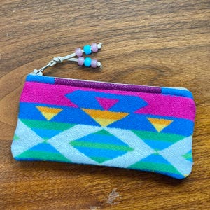 May include: A rectangular zippered pouch featuring a vibrant woven pattern of hot pink, bright blue, green, yellow, and white geometric shapes. The zipper pull is accented with two light pink and two bright blue plastic beads on a cord.