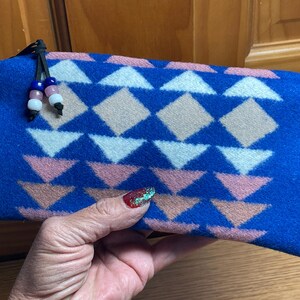 May include: A blue rectangular pouch with a geometric pattern of white, pink, and tan shapes. The pouch has a black zipper pull with decorative beads. The pouch is held in a hand.
