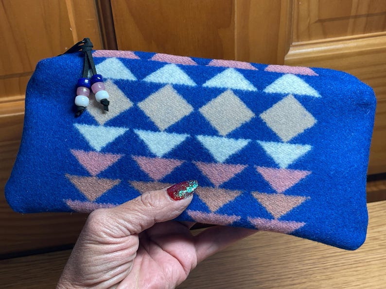 May include: A blue rectangular pouch with a geometric pattern of white, pink, and tan shapes. The pouch has a black zipper pull with decorative beads. The pouch is held in a hand.