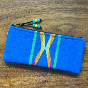 May include: A rectangular blue fabric pencil case featuring a central woven pattern of intersecting orange, yellow, and green lines. The zipper pull is adorned with two black cords, each tipped with a pair of small, bright blue and yellow plastic beads.