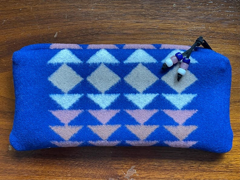 May include: A blue rectangular pencil case with a geometric pattern of white, pink, and tan diamonds and triangles. The zipper pull has decorative beads. The case is made of a soft, textured material.