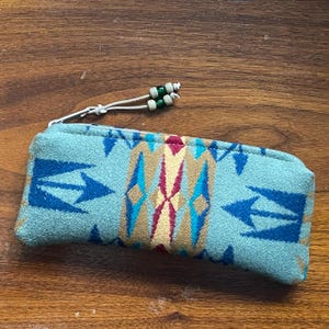 May include: A rectangular, turquoise fabric pencil case with a Southwestern pattern in blue, gold, and maroon. The case has a white drawstring with green and tan beads. The case is on a wooden surface.