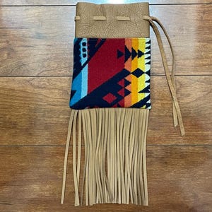 May include: A tan leather pouch with a colorful geometric design. The pouch has a drawstring closure and long fringe at the bottom. The design includes red, blue, yellow, and black patterns.