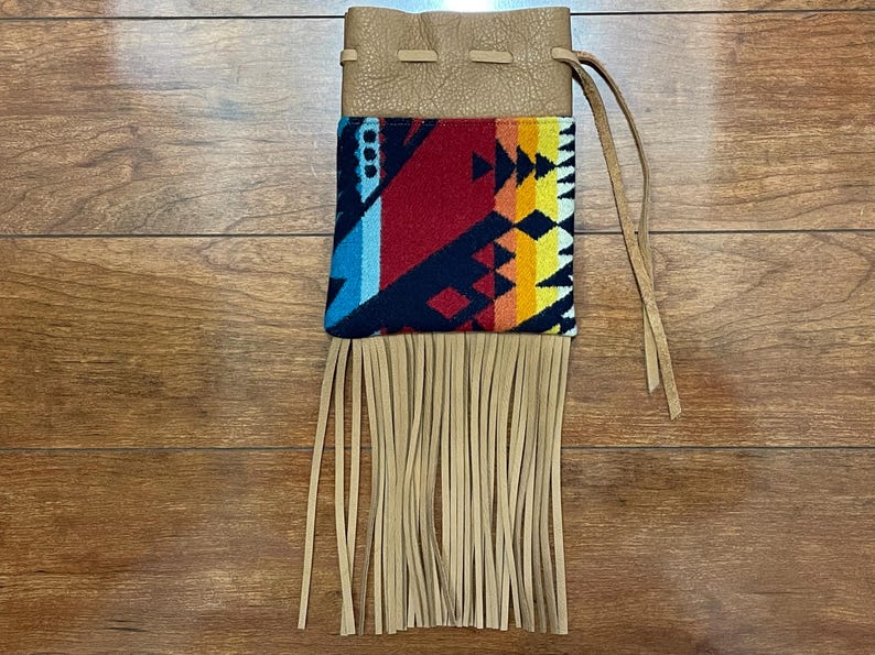 May include: A tan leather pouch with a colorful geometric design. The pouch has a drawstring closure and long fringe at the bottom. The design includes red, blue, yellow, and black patterns.