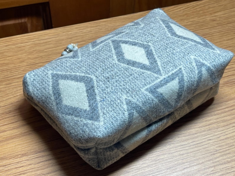 Puede incluir: Un art&iacute;culo de tela rectangular y plegado con un patr&oacute;n geom&eacute;trico de diamantes en tonos grises y blanquecinos. El art&iacute;culo parece ser un embrague o una bolsa peque&ntilde;a, con un detalle de cuentas en un extremo. La tela tiene una apariencia texturizada.