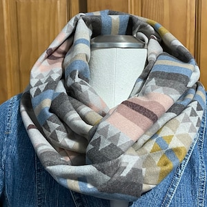 May include: A soft, patterned infinity scarf in shades of gray, blue, pink, and yellow. The scarf features a geometric design with triangles and stripes. The scarf is wrapped around a mannequin and worn over a denim jacket.