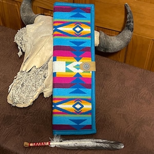 Puede incluir: Un artículo textil rectangular y colorido con un diseño del suroeste. El artículo presenta un borde turquesa y un patrón de formas geométricas azules, rosas, amarillas y blancas. Una correa de cuero con un botón plateado asegura el artículo. Una pluma y una calavera también están en la imagen.