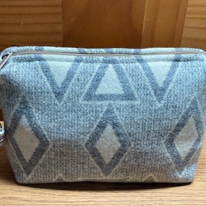 Puede incluir: Peque&ntilde;o neceser rectangular con un estampado geom&eacute;trico de diamantes y tri&aacute;ngulos en azul claro y blanco. El neceser tiene cierre de cremallera y un peque&ntilde;o tirador decorativo. El neceser est&aacute; hecho de tela suave y texturizada.