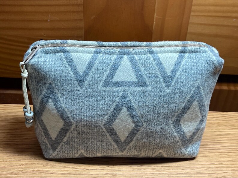 Puede incluir: Peque&ntilde;o neceser rectangular con un estampado geom&eacute;trico de diamantes y tri&aacute;ngulos en azul claro y blanco. El neceser tiene cierre de cremallera y un peque&ntilde;o tirador decorativo. El neceser est&aacute; hecho de tela suave y texturizada.