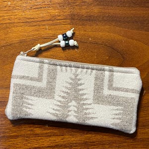 May include: A rectangular pencil case with a cream and beige geometric pattern. The zip pull has a small beaded accent with white and dark green beads. The case is made of a soft, textured fabric.