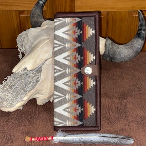 Puede incluir: Una cartera rectangular marrón con un patrón geométrico en gris, blanco, rojo y beige. La cartera tiene una correa de cuero con cierre de botón. Una pluma decorativa y una calavera con cuernos también están en la imagen.