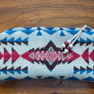 May include: A rectangular, soft-sided pouch with a geometric pattern in white, blue, and red. The design features triangles and diamond shapes. The pouch has a white drawstring with red and white beads.