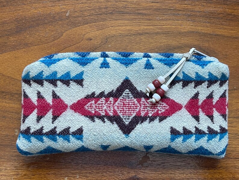 May include: A rectangular, soft-sided pouch with a geometric pattern in white, blue, and red. The design features triangles and diamond shapes. The pouch has a white drawstring with red and white beads.