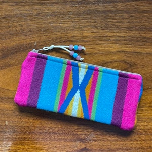 May include: A rectangular zippered pouch featuring a vibrant multi-colored striped pattern in shades of hot pink, purple, bright blue, green, yellow, and white. The zipper pull is accented with two white cords decorated with light blue and pink beads.