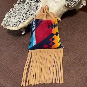May include: A small, colorful pouch with a geometric pattern in red, blue, and yellow. The pouch has a tan leather top and long fringe. The drawstring closure is also tan leather.
