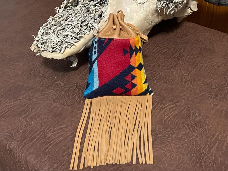 May include: A small, colorful pouch with a geometric pattern in red, blue, and yellow. The pouch has a tan leather top and long fringe. The drawstring closure is also tan leather.