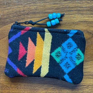 May include: A small, rectangular black coin purse with a colorful geometric pattern in shades of red, orange, yellow, blue, and purple. The purse has a black zipper and a beaded pull with turquoise and blue beads.
