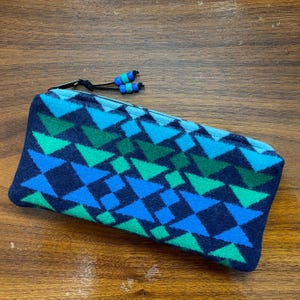 May include: A rectangular pencil case with a geometric pattern of blue and green triangles and diamonds on a navy blue background. The zipper pull has blue and green beads.