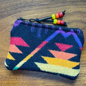 May include: A small, rectangular black zippered pouch with a colorful geometric pattern in shades of yellow, orange, pink, purple, and blue. The zipper pull features a leather cord with red, orange, and yellow beads.