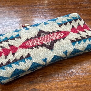 May include: Rectangular, patterned case with a cream background and geometric designs in teal, burgundy, and brown. The case has a woven texture and a wooden bead closure. The case is likely for glasses or small items.