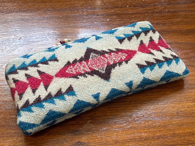 May include: Rectangular, patterned case with a cream background and geometric designs in teal, burgundy, and brown. The case has a woven texture and a wooden bead closure. The case is likely for glasses or small items.