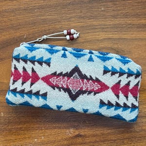 May include: A rectangular, zippered pouch with a geometric pattern in shades of blue, pink, and black. The pouch has a white background and a beaded pull. The pouch is approximately 8 inches long.