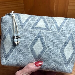 Puede incluir: Un estuche rectangular con un patr&oacute;n geom&eacute;trico azul claro y blanco de diamantes y tri&aacute;ngulos. El estuche tiene un cierre de cremallera y un cord&oacute;n color crema con cuentas decorativas. La tela es suave.